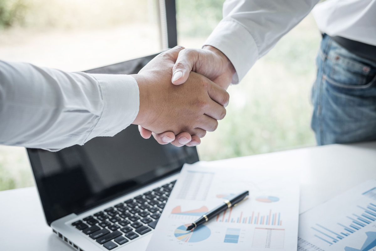 A deal being made between an energy broker and a customer. Shaking hands over a laptop and paper showing graphs.