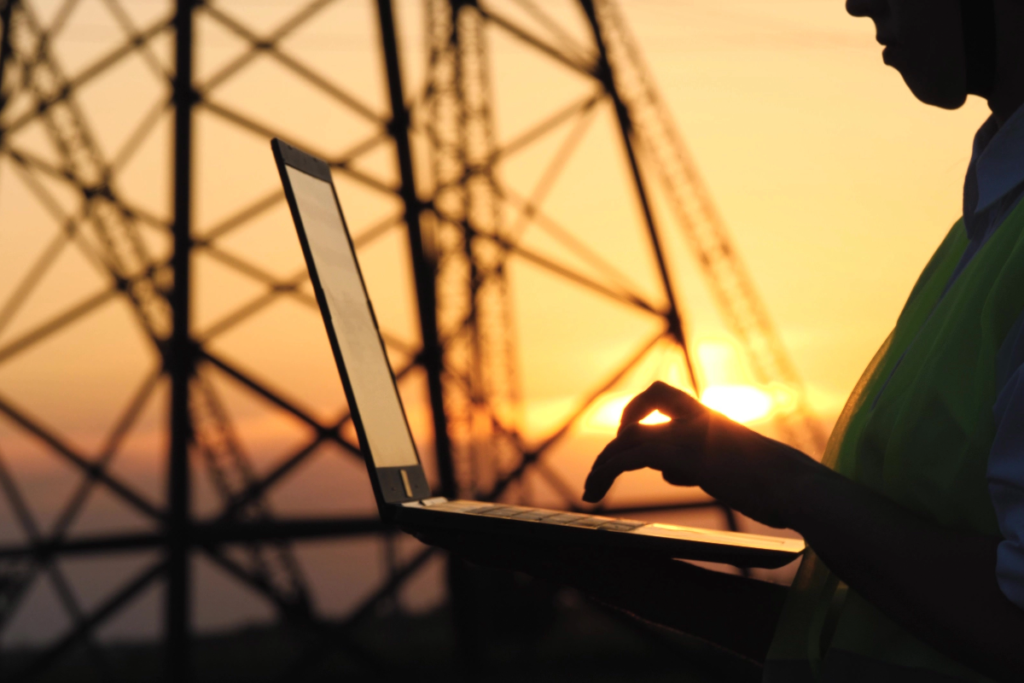 Someone in hi-vis using a laptop in front of an electricity pylon during sunset